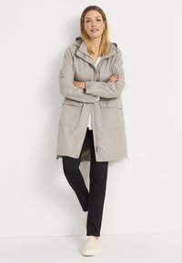 Lichtgrijze waterdichte parka met een capuchon, voorzien van een ritssluiting aan de voorkant, twee grote zakken en een losse pasvorm. G gecombineerd met zwarte broeken en beige schoenen.