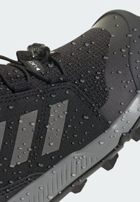 Black athletic shoe with textured upper, featuring water droplets. Gray accents and three stripes on the side, rugged sole for traction.