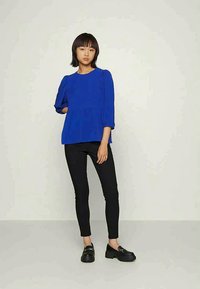 Top azul texturizado com mangas bufantes e cintura franzida, combinado com calças pretas ajustadas e sapatos pretos de deslizar.