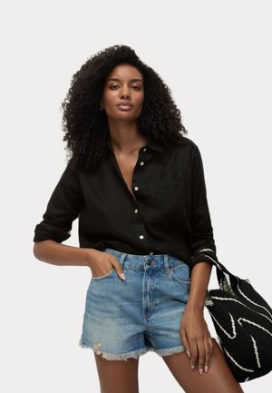 Chemise noire à boutons en lin avec des manches retroussées, associée à un short en denim bleu clair avec des ourlets effilochés et des détails usés.