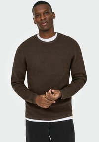 Pull en maille marron avec un design côtelé, encolure ronde et manches longues, porté par-dessus une chemise blanche ; élégant et ajusté.