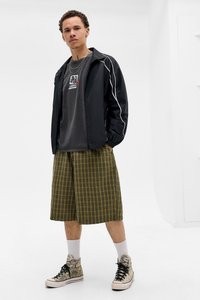 Fekete, könnyű dzseki fehér csíkokkal, szürke póló logóval, zöld kockás sort és mintás magas szárú sportcipő.