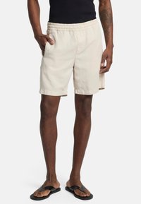Beige katoenen shorts met een elastische tailleband, zijzakken en omgeslagen zoom, gecombineerd met zwarte slippers op een model tegen een witte achtergrond.