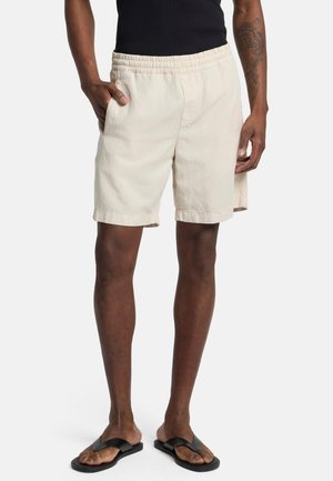 Beige Baumwollshorts mit einem elastischen Bund, seitlichen Taschen und aufgerollten Säumen, kombiniert mit schwarzen Flip-Flops an einem Modell vor einem weißen Hintergrund.