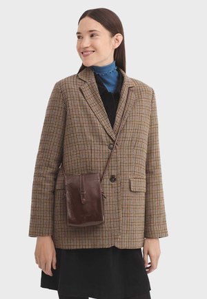 Braun karierter Blazer mit zwei Knöpfen und Taschen, getragen über einem blauen Rollkragenpullover. Eine kleine braune Ledertasche hängt seitlich.