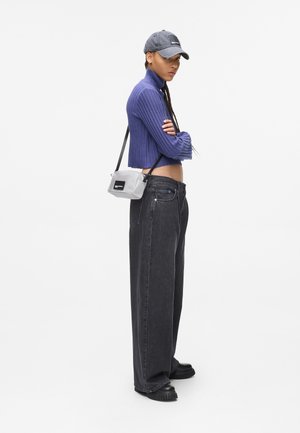 Giovane in piedi con le braccia incrociate, indossando un maglione corto viola, un cappello grigio, jeans neri larghi, scarpe nere e una borsa a tracolla grigia.