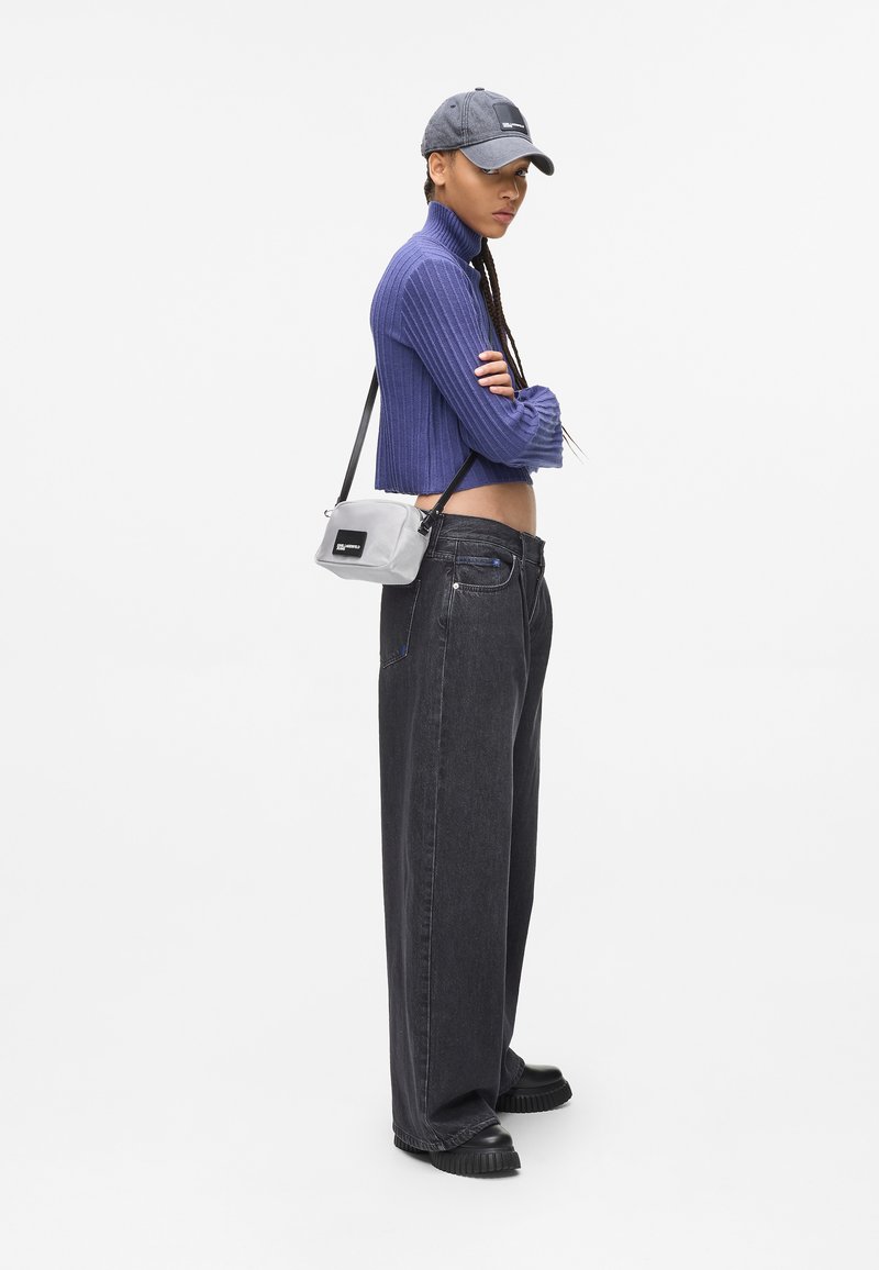 Giovane in piedi con le braccia incrociate, indossando un maglione corto viola, un cappello grigio, jeans neri larghi, scarpe nere e una borsa a tracolla grigia.