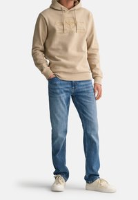 Sudadera con capucha beige con logo bordado, cordón ajustable y puños acanalados. Combinada con vaqueros de mezclilla azul claro y zapatillas crema.