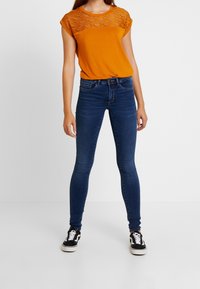 Frau in einem orangefarbenen, kurzärmeligen Oberteil mit Spitze, dunklen, enganliegenden Jeans und schwarzen Sneakers mit weißen Schnürsenkeln, die vor einem weißen Hintergrund steht.