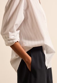 Wit gestreept shirt met opgerolde mouwen, semi-loose fit. Gecombineerd met donkere broeken. Verticale strepen en een gladde stofstructuur zijn opvallend.
