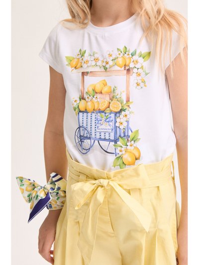Κορίτσι με λευκό μπλουζάκι με εκτύπωση καροτσιού με λεμόνια και λουλούδια, κίτρινο παντελόνι paperbag με φιόγκο και μαντήλι με σχέδιο λεμονιού δεμένο στον καρπό.