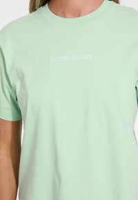 Minttuvihreä puuvillapaita, jossa on pyöreä kaula-aukko ja lyhyet hihat. Paidassa on rintataskussa brodeerattu valkoinen "GUESS JEANS" -logo.