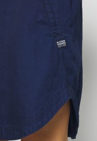 Tummansininen G-Star RAW Originals farkuhame, jossa on näkyvä merkkilogo tikattua taskua lähellä ja sivuriiputus, joka paljastaa ihoa.
