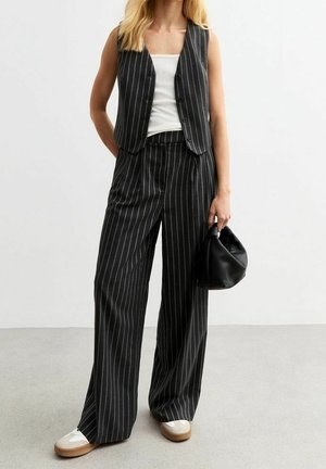 Frau trägt anthrazitfarbene Nadelstreifen-Weitbein-Hose und passende Weste über weißem Top, hält eine kleine schwarze Handtasche und trägt weiße Sneaker.