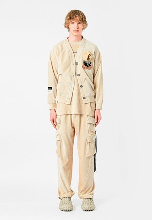 Chaqueta de pana beige con botones y un parche, combinada con pantalones cargo a juego que tienen bolsillos. Llevada con zapatos texturizados beige.