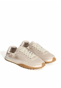 Zapatillas sintéticas en color beige con un acabado brillante, que cuentan con cordones redondos, puños acolchados y una suela de goma texturizada. Logotipo de la marca mostrado en el costado.