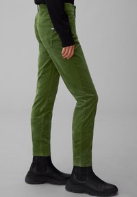 Persona che indossa pantaloni in corda verde e stivaletti neri, in piedi contro uno sfondo grigio uniforme.