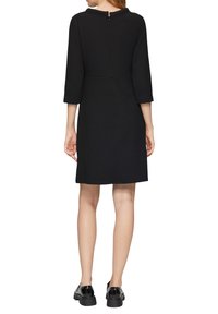 Robe noire avec un motif texturé, des manches trois-quarts et une fermeture éclair au dos. Associée à des chaussures noires à talons épais.