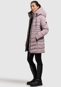Abrigo de plumas rosa claro con capucha, costuras verticales, bolsillos con cremallera y un diseño elegante. Combinado con leggings negros y botas altas negras.