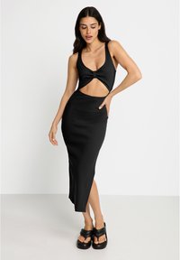 Naisso wearing a black sleeveless cutout dress and black platform sandals, posing with one hand on hip and the other relaxed.