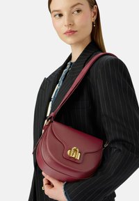 Žena v černém pruhovaném saku nese hnědou koženou přes rameno tašku s zlatým zapínáním.