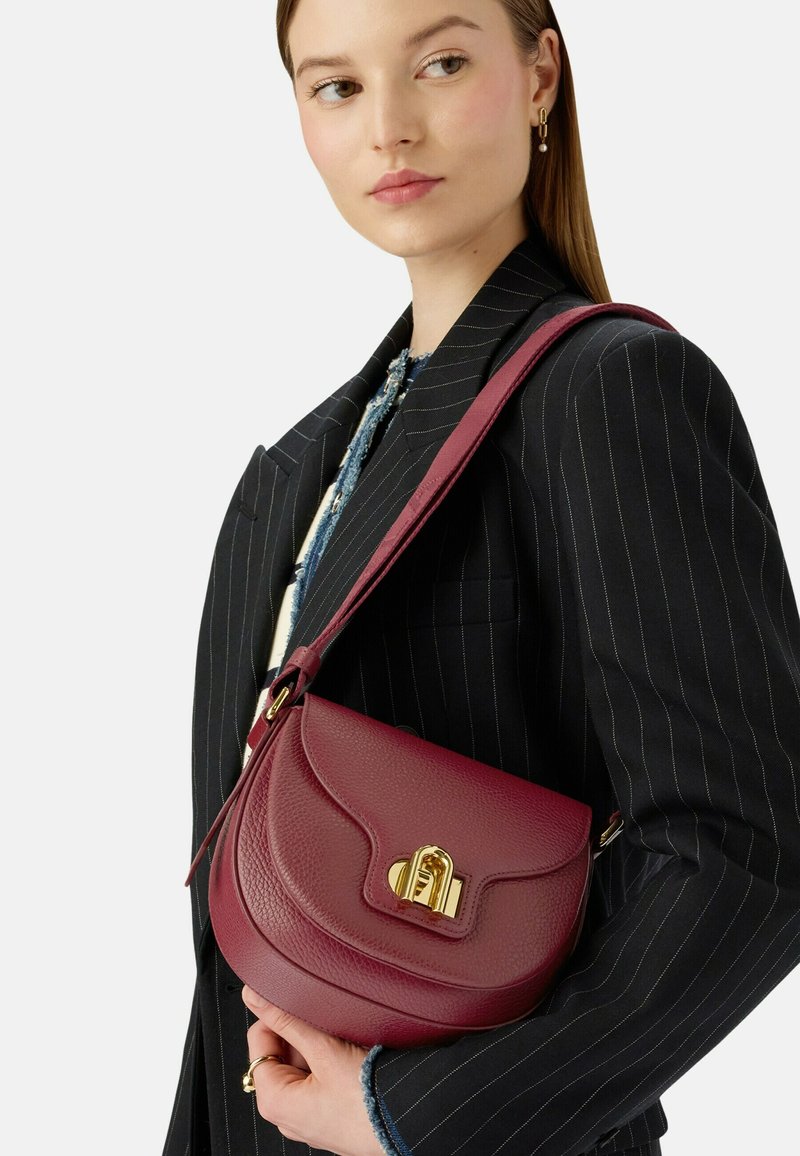 Žena v černém pruhovaném saku nese hnědou koženou přes rameno tašku s zlatým zapínáním.