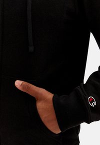 Felpa nera con cappuccio, dotata di cordoncino, tasca frontale e polsini a coste, con un piccolo logo di accentuazione sulla manica. Tessuto dalla consistenza morbida.