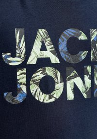 Κείμενο "JACK JONES" με γράμματα γεμάτα με πράσινα και μπλε τροπικά φυλλαράκια πάνω σε υπόβαθρο από σκούρο ναυτικό ύφασμα.