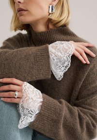 Pull en laine marron avec design côtelé, featuring des poignets en dentelle blanche. Bague argentée portée à la main sur un jean denim bleu clair. Texture détaillée visible.