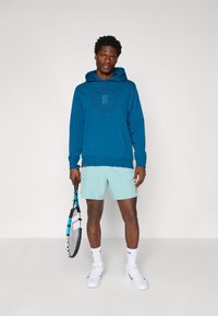 Blå hættetrøje med en frontlomme, snor i hætten og logo. Parret med lyseblå shorts, holder en tennisracket. Hvide sneakers og sokker.