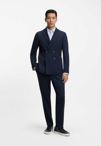 Laivastonsininen kaksirivinen bleiseri, jossa kaksi taskua, yhdistetty samansävyisiin housuihin ja mustiin lenkkareihin. Sileä kangas, mittatilaustyönä tehty istuvuus, siisti muotoilu.