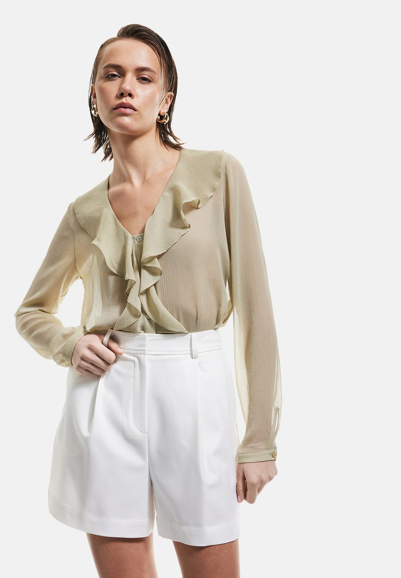 Blouse verte clair à volants avec manches longues et fermeture à boutons, associée à un short blanc ajusté avec des plis et des passants de ceinture.