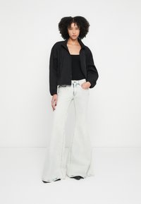 Must zip-up bomberjakk, kehastunud must topp, helesinised lõhestatud säärega teksapüksid, modell seisab käed puusadel, loomulikud lokkis juuksed.