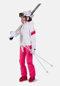 Skioutfit met een witte jas met rode en gele accenten, roze skibroek, witte handschoenen, rode skischoenen en skiëquipment.