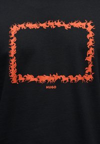 Μαύρο ύφασμα με ορθογώνιο περίγραμμα από πορτοκαλί σιλουέτες αλόγου και αναβάτη που περιβάλλουν το όνομα της μάρκας "HUGO" στο κάτω κέντρο.