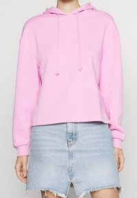 Sudadera con capucha rosa de corte holgado, con cordón en la capucha y puños acanalados, combinada con una falda vaquera azul claro con dobladillo deshilachado.