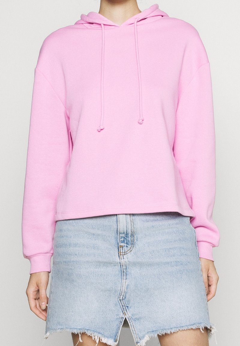 Sudadera con capucha rosa de corte holgado, con cordón en la capucha y puños acanalados, combinada con una falda vaquera azul claro con dobladillo deshilachado.