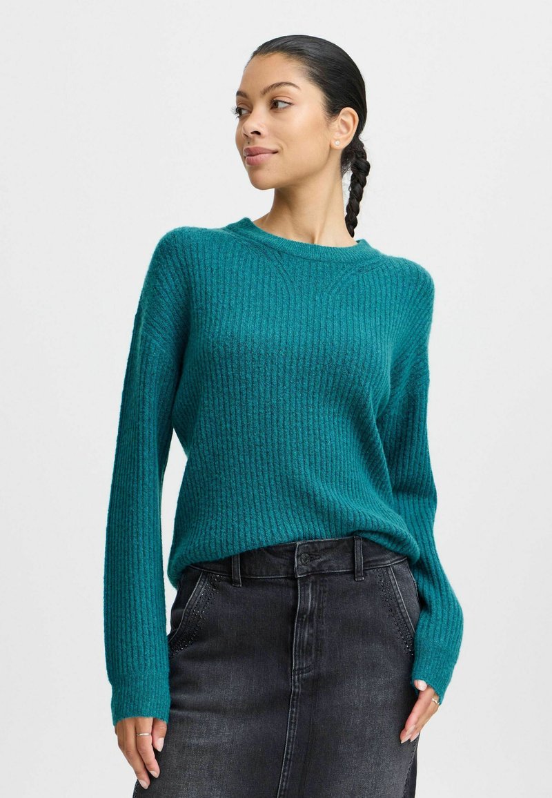 Pull en tricot teal avec une texture côtelée, un col rond et des manches longues, associé à une jupe en denim noire. Design lisse et décontracté.