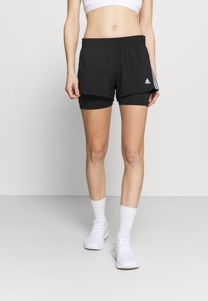 Vrouw draagt zwarte gelaagde sportshorts, witte sportbeha, witte sokken en witte hardloopschoenen, staand op een effen achtergrond.