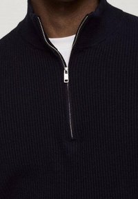 Marineblauer gerippter Pullover mit hohem Kragen und halbem Reißverschluss. Besitzt silberne Reißverschlussbeschläge und ein strukturiertes Strickmuster.