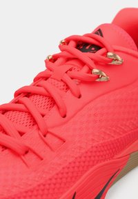 Φωτεινό ροζ αθλητικό παπούτσι με υφή στο επάνω μέρος, πάνελ από δίχτυ, χρυσές λεπτομέρειες στα κορδόνια και μαύρο λογότυπο στο πλάι.