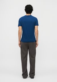 Άνδρας με σγουρά μαύρα μαλλιά, ντυμένος με μπλε κοντομάνικο πουκάμισο και φαρδιά σκούρα γκρι παντελόνια, στέκεται με την πλάτη προς τα εμπρός σε απλό φόντο.