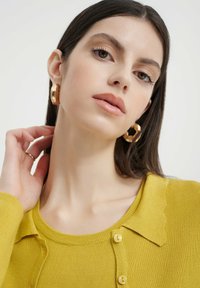 Gelber, gerippter Cardigan mit einem gewellten Kragen und kleinen Knöpfen. Goldene Creolen sind sichtbar. Der Hautton wirkt gleichmäßig mit subtiler Make-up.