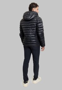 Giubbotto imbottito nero con cappuccio, caratterizzato da un design trapuntato e una texture lucida. Indossato sopra pantaloni neri e sneakers di colore chiaro.