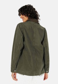 Veste en coton vert olive avec un col côtelé, des détails de boutons et une coupe décontractée. Coutures visibles et léger brillant sur le tissu.