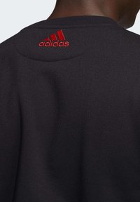 Vue arrière d'une personne portant un vêtement noir avec le logo Adidas rouge brodé au niveau de l'encolure arrière.