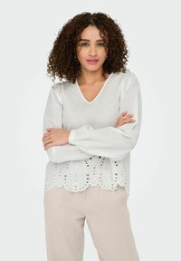 Weiße Bluse mit langen Ärmeln, V-Ausschnitt und Spitzenbesatz mit ausgeschnittenen Mustern. Leichter Stoff mit einem leicht durchsichtigen Textur.