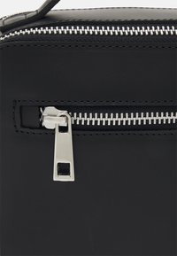 Pochette en simili cuir noir avec fermoir à glissière en argent, présentant une forme rectangulaire et une surface texturée entourant la fermeture.