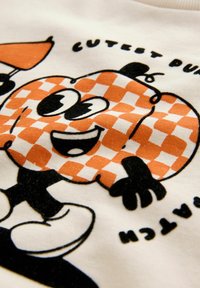 Weißer Stoff mit einem Cartoon-Kürbis, der ein karottenartiges oranges Muster aufweist, große Augen und einen lächelnden Gesichtsausdruck hat, umgeben von schwarzem Text.