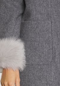 Manche d'un manteau en laine grise avec un poignet en fourrure gris clair et une poche avant apparente, main reposant à côté du manteau.